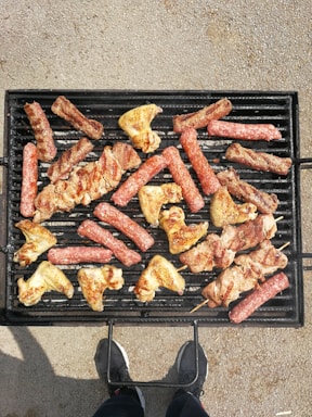 Various types of meat, including sausages, chicken wings, and skewered pieces, are being grilled on a black barbecue grill. The grill is placed on a textured ground surface and there's a person standing in front of it, visible only from the waist down and wearing dark shoes.