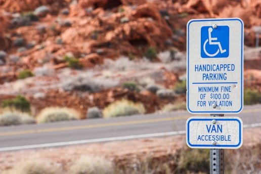 A parking sign designating handicapped parking with a minimum fine for violation. The sign also indicates that the space is van accessible. It is set against a background of red rocky terrain and roadside vegetation.
