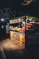 A juice stand in a dimly lit setting, featuring an array of fresh fruits like strawberries, watermelons, and lemons. The wooden stall has a rustic look with its name 'Juicebox London' prominently displayed. The stall is adorned with small chalkboard menus, and there is a person working inside the stand.