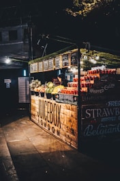A juice stand in a dimly lit setting, featuring an array of fresh fruits like strawberries, watermelons, and lemons. The wooden stall has a rustic look with its name 'Juicebox London' prominently displayed. The stall is adorned with small chalkboard menus, and there is a person working inside the stand.
