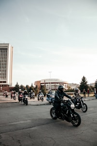 A group of motorcyclists wearing helmets and protective gear ride their bikes on a city street. The sky is clear, and there are trees and modern buildings in the background. Bystanders are visible on the sidewalk, observing the riders.