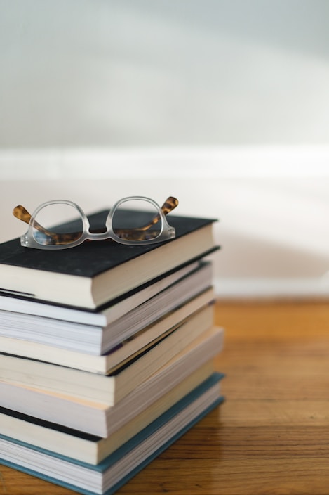 A stack of books is neatly piled on a wooden surface, with a pair of reading glasses resting on the top book. The scene is set against a softly lit background, creating a calm and inviting atmosphere.
