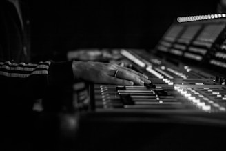 A close-up view of a person's hand adjusting controls on a professional audio mixing console. The scene is captured in black and white, highlighting the intricate details of the equipment and the focused action.