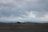A downed aircraft lies on a barren, rocky landscape, while a group of people explore the site. The sky is overcast with heavy clouds, creating a dramatic backdrop. The ocean can be seen in the distance, contributing to the desolate and isolated feeling of the scene.