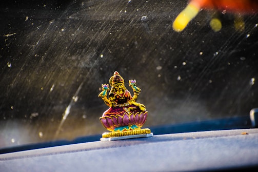 A small, colorful statue of a deity sitting on a lotus flower is placed on a car dashboard. The deity is holding objects in both hands. The background appears to be a car windshield with dust and streaks visible.