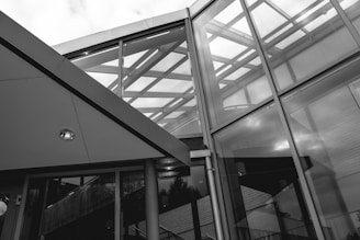 A modern architectural structure featuring a combination of glass and metal elements, showcasing geometric and angular lines. The image captures reflections and transparency of glass panels, revealing an intricate framework and a glimpse of the sky through the roof.