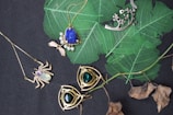 An assortment of jewelry is arranged on a dark surface with vibrant green leaves. The collection includes necklaces with intricate designs featuring gemstones. Notable pieces include a spider-shaped pendant with a white gemstone and a necklace with a large blue stone surrounded by smaller pearls. Another item is a silver pendant with branch-like elements. The background contrasts with dried leaves adding a natural element.