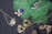 An assortment of jewelry is arranged on a dark surface with vibrant green leaves. The collection includes necklaces with intricate designs featuring gemstones. Notable pieces include a spider-shaped pendant with a white gemstone and a necklace with a large blue stone surrounded by smaller pearls. Another item is a silver pendant with branch-like elements. The background contrasts with dried leaves adding a natural element.