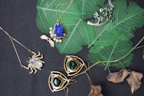 An assortment of jewelry is arranged on a dark surface with vibrant green leaves. The collection includes necklaces with intricate designs featuring gemstones. Notable pieces include a spider-shaped pendant with a white gemstone and a necklace with a large blue stone surrounded by smaller pearls. Another item is a silver pendant with branch-like elements. The background contrasts with dried leaves adding a natural element.