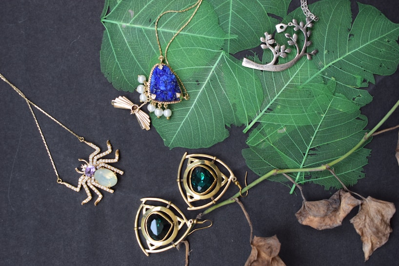 An assortment of jewelry is arranged on a dark surface with vibrant green leaves. The collection includes necklaces with intricate designs featuring gemstones. Notable pieces include a spider-shaped pendant with a white gemstone and a necklace with a large blue stone surrounded by smaller pearls. Another item is a silver pendant with branch-like elements. The background contrasts with dried leaves adding a natural element.