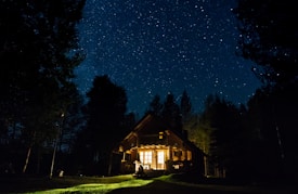 A cozy cabin warmly illuminated with lights, nestled amidst tall trees under a stunning, star-filled night sky.