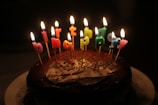 A chocolate cake topped with lit candles spelling out 'happy birthday'. The candles are colorful, and the backdrop is dark, making the flames stand out brightly against the cake.