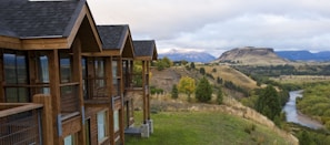 A wooden lodge-style building with multiple gabled roofs stands on the edge of a grassy hill, overlooking a scenic landscape. In the background, a river winds through a lush green valley surrounded by rolling hills and distant mountains under a cloudy sky.