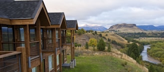 A wooden lodge-style building with multiple gabled roofs stands on the edge of a grassy hill, overlooking a scenic landscape. In the background, a river winds through a lush green valley surrounded by rolling hills and distant mountains under a cloudy sky.