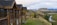 A wooden lodge-style building with multiple gabled roofs stands on the edge of a grassy hill, overlooking a scenic landscape. In the background, a river winds through a lush green valley surrounded by rolling hills and distant mountains under a cloudy sky.