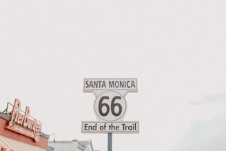 A classic Route 66 sign is prominently displayed, reading 'Santa Monica 66 End of the Trail.' Nearby, part of a building with a sign for 'Pier Burger' is visible against a light, cloudy sky.