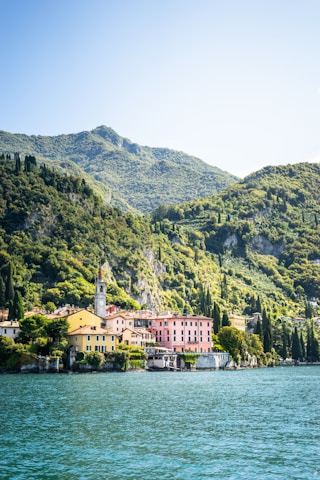 Colorful buildings with pastel facades are nestled along a picturesque lakeside, backdropped by lush green hills covered with trees. A small bell tower rises above the buildings, adding a touch of historic charm. The clear blue water in the foreground complements the vibrant greenery of the landscape.