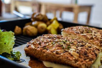 A close-up of a sandwich with a multi-seed crust and melted cheese, accompanied by roasted potatoes and a side of fresh green lettuce. The scene is set on a dark plate, with a blurred background of a wooden table and blurred dining area.