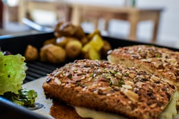 A close-up of a sandwich with a multi-seed crust and melted cheese, accompanied by roasted potatoes and a side of fresh green lettuce. The scene is set on a dark plate, with a blurred background of a wooden table and blurred dining area.
