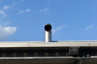 A metal vent pipe protrudes from the roof of a building against a clear blue sky with a few scattered clouds. The roof features an industrial design with visible beams and circular openings, and the vent pipe adds to the utilitarian feel.