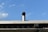 A metal vent pipe protrudes from the roof of a building against a clear blue sky with a few scattered clouds. The roof features an industrial design with visible beams and circular openings, and the vent pipe adds to the utilitarian feel.