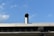 A metal vent pipe protrudes from the roof of a building against a clear blue sky with a few scattered clouds. The roof features an industrial design with visible beams and circular openings, and the vent pipe adds to the utilitarian feel.