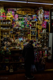 A crowded artisan shop filled with vibrant colors and a variety of small items ranging from toys to handcrafted goods. The shop is tightly packed with shelves displaying colorful masks, traditional musical instruments, mugs, and woven crafts. A person stands in front of the shop browsing or purchasing an item, holding a plastic bag.
