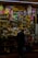 A crowded artisan shop filled with vibrant colors and a variety of small items ranging from toys to handcrafted goods. The shop is tightly packed with shelves displaying colorful masks, traditional musical instruments, mugs, and woven crafts. A person stands in front of the shop browsing or purchasing an item, holding a plastic bag.