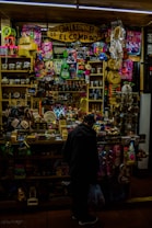 A crowded artisan shop filled with vibrant colors and a variety of small items ranging from toys to handcrafted goods. The shop is tightly packed with shelves displaying colorful masks, traditional musical instruments, mugs, and woven crafts. A person stands in front of the shop browsing or purchasing an item, holding a plastic bag.