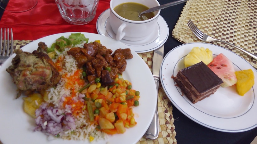 A variety of food is arranged on white plates, including rice mixed with vegetables, grilled meat, and a rich stew. Accompanying the meal are a cup of soup and a dessert plate featuring chocolate cake and slices of fresh fruit such as watermelon and pineapple. The table setting includes a glass, cutlery, and a woven placemat.