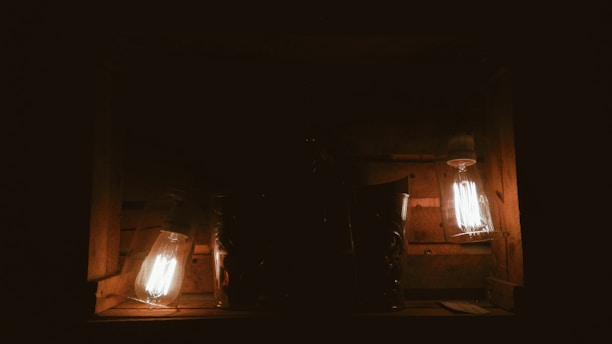 A dark wooden box with two illuminated vintage-style light bulbs emitting a warm glow. The light softly highlights the textured wooden surface and a metallic vase-like object.