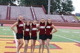 A group of five cheerleaders stand in a row on a sports field. They are wearing matching maroon tops with the letters LBMS and black skirts. The background consists of empty bleachers and trees.
