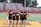 A group of five cheerleaders stand in a row on a sports field. They are wearing matching maroon tops with the letters LBMS and black skirts. The background consists of empty bleachers and trees.