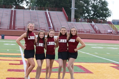 A group of five cheerleaders stand in a row on a sports field. They are wearing matching maroon tops with the letters LBMS and black skirts. The background consists of empty bleachers and trees.
