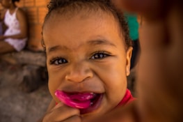 A close-up of a young child with expressive eyes holding a pink pacifier in their mouth. The background is slightly blurred, showing another person sitting, wearing a white and pink outfit against a brick wall.