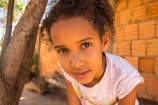 A young girl with curly hair is looking directly at the camera, wearing a light-colored shirt. She is outdoors beside a tree with a brick wall in the background, illuminated by natural light.