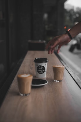 A hand reaches for a takeaway coffee cup on a long wooden table. The cup features the words 'Say Something' in a handwritten style. Two glasses of latte sit alongside the cup, with one placed on a black saucer. The setting appears to be a cafe with a blurred street view visible in the background.
