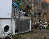 A scene depicting an outdoor urban environment with discarded household appliances, including a washing machine and a refrigerator, placed against a graffiti-covered stone wall. The area is cluttered with debris, such as pieces of wood, metal, and other trash. Sparse greenery grows amidst the concrete and rubble.