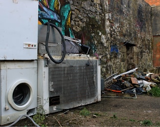 A scene depicting an outdoor urban environment with discarded household appliances, including a washing machine and a refrigerator, placed against a graffiti-covered stone wall. The area is cluttered with debris, such as pieces of wood, metal, and other trash. Sparse greenery grows amidst the concrete and rubble.