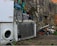 A scene depicting an outdoor urban environment with discarded household appliances, including a washing machine and a refrigerator, placed against a graffiti-covered stone wall. The area is cluttered with debris, such as pieces of wood, metal, and other trash. Sparse greenery grows amidst the concrete and rubble.