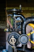 A close-up view of a vintage machine with visible gears and metal tools attached to it. The machine has an industrial, mechanical appearance with engraved text on the metal parts. It appears to be designed for a specific manufacturing or mechanical process.