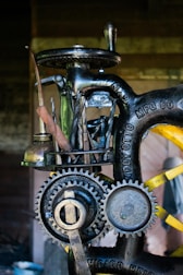 A close-up view of a vintage machine with visible gears and metal tools attached to it. The machine has an industrial, mechanical appearance with engraved text on the metal parts. It appears to be designed for a specific manufacturing or mechanical process.
