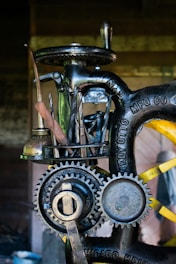 A close-up view of a vintage machine with visible gears and metal tools attached to it. The machine has an industrial, mechanical appearance with engraved text on the metal parts. It appears to be designed for a specific manufacturing or mechanical process.