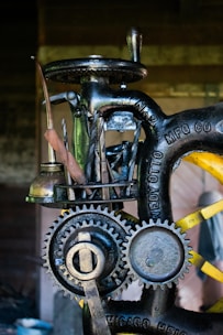 A close-up view of a vintage machine with visible gears and metal tools attached to it. The machine has an industrial, mechanical appearance with engraved text on the metal parts. It appears to be designed for a specific manufacturing or mechanical process.