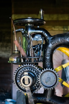 A close-up view of a vintage machine with visible gears and metal tools attached to it. The machine has an industrial, mechanical appearance with engraved text on the metal parts. It appears to be designed for a specific manufacturing or mechanical process.