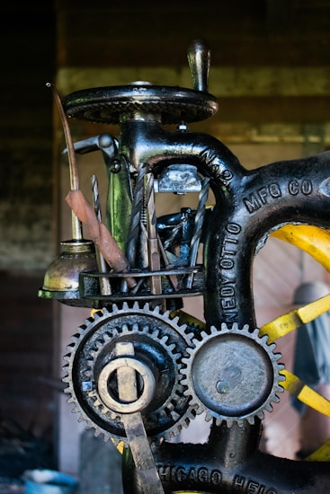A close-up view of a vintage machine with visible gears and metal tools attached to it. The machine has an industrial, mechanical appearance with engraved text on the metal parts. It appears to be designed for a specific manufacturing or mechanical process.
