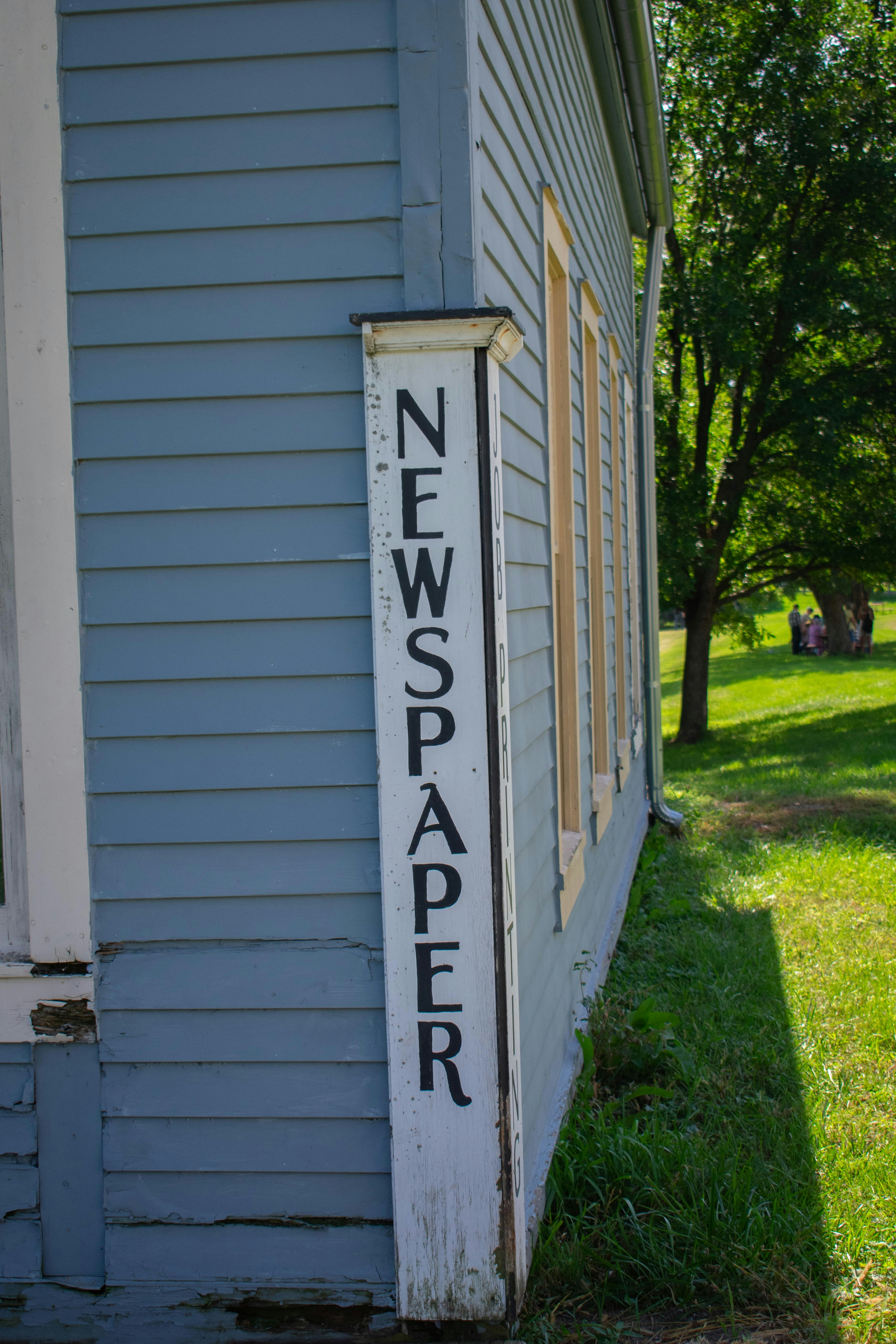 A vintage wooden building with light blue siding features a sign with the word 'NEWSPAPER' in large black letters painted on a white vertical board. The structure is set against a backdrop of lush green grass and trees, with sunlight casting shadows on the building.