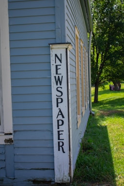 A vintage wooden building with light blue siding features a sign with the word 'NEWSPAPER' in large black letters painted on a white vertical board. The structure is set against a backdrop of lush green grass and trees, with sunlight casting shadows on the building.