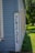 A vintage wooden building with light blue siding features a sign with the word 'NEWSPAPER' in large black letters painted on a white vertical board. The structure is set against a backdrop of lush green grass and trees, with sunlight casting shadows on the building.