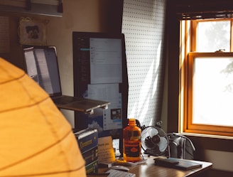 A cozy workspace with sunlight streaming through a window onto a desk. The desk features a laptop on a stand, a large monitor displaying text and images, a small fan, an orange water bottle, and some stationery items. Books are stacked on one side, and a thank-you note is visible on the desk.